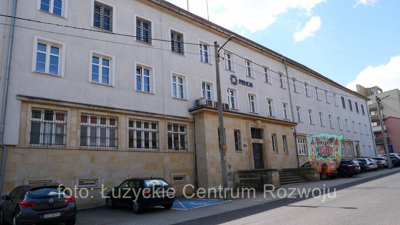 duży dwupiętrowy budynek z napisem POLICJA, przed budynkiem parking z samochodami osobowymi